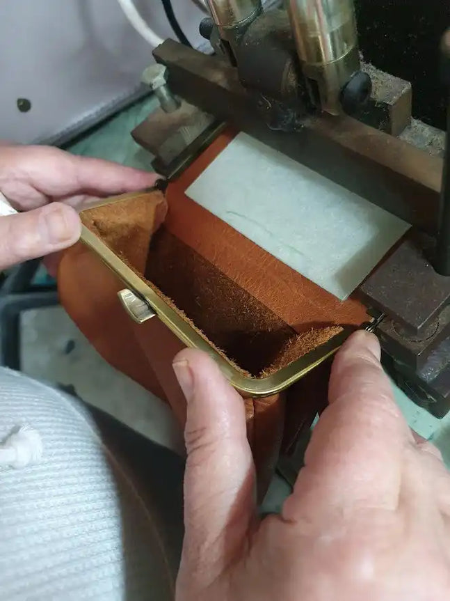 Hands using a leather working tool to stitch or work on a brown leather piece.