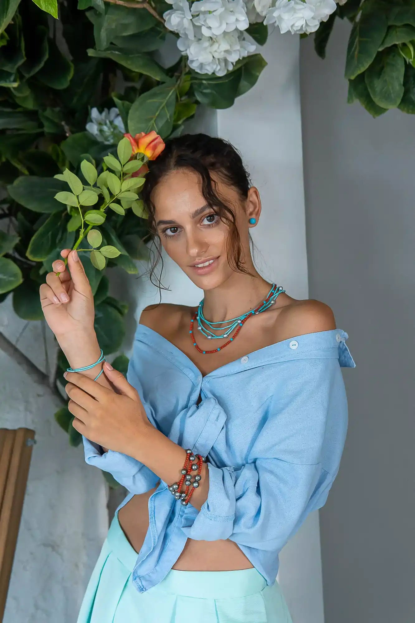 A person wearing a light blue off-shoulder top and turquoise jewelry poses with a flower.