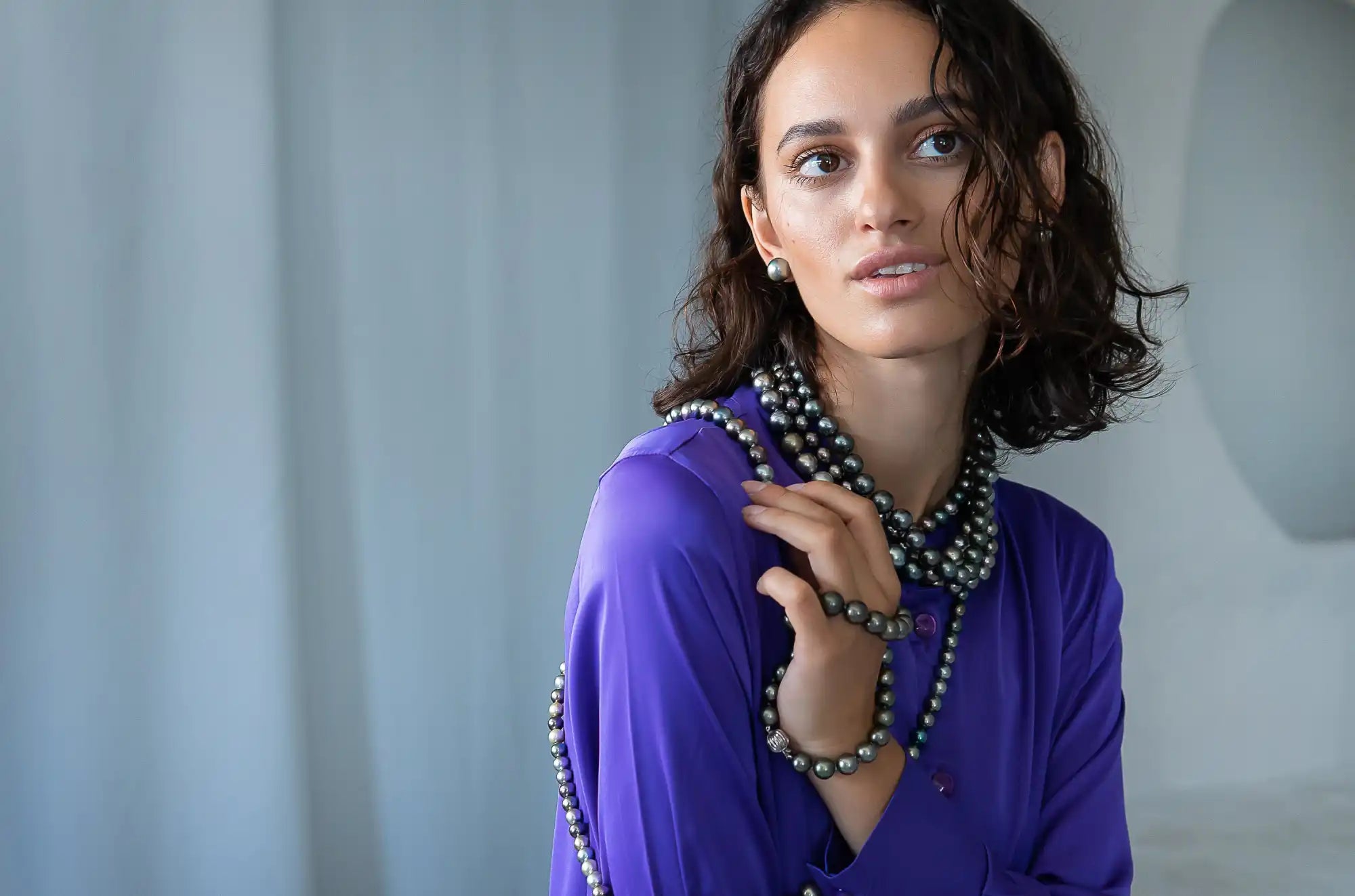 A woman with dark, curly hair wearing a vibrant purple top and multiple strands of dark pearls as necklaces and bracelets.